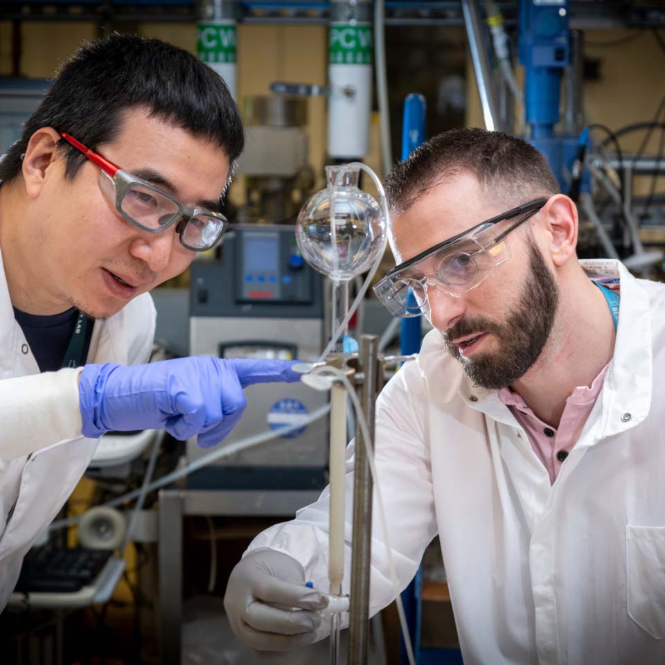 Two student chemists wearing protective goggles, gloves and lab coats enact an experiment