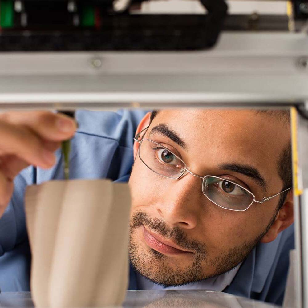 A male student wearing glasses adjusts a 3D printer with a partially finished curvy beige vase print