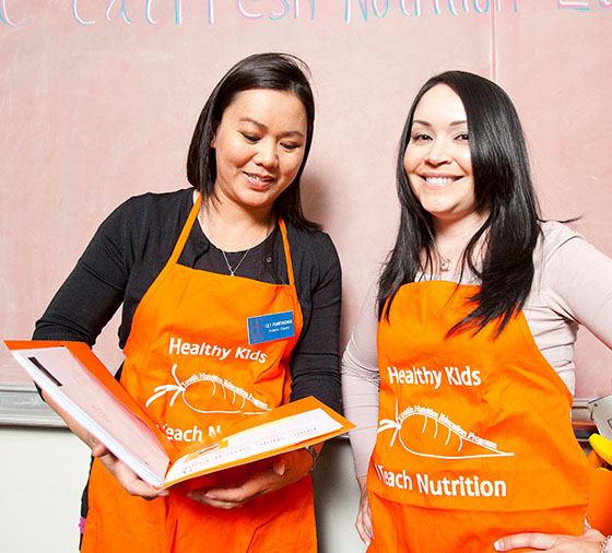 Two women wearing orange aprons that read 'Healthy Kids - I Teach Nutrition'. One holds an orange folder open.