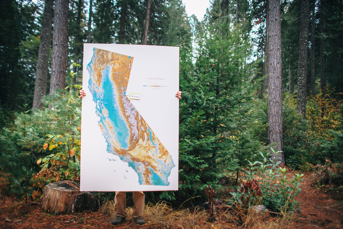 A person holds a ~6-foot tall topographic map of California, standing in a forest.